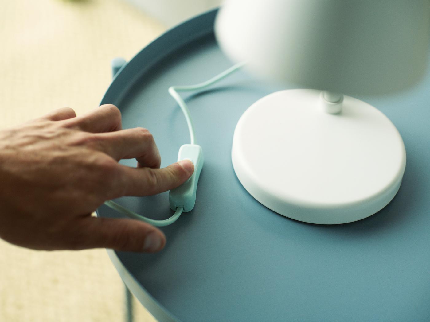 Une main actionne l’interrupteur d’une lampe de table blanche posée sur une table bleu clair.