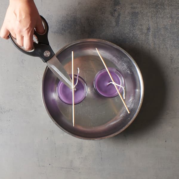 Une main coupe la mèche de deux bougies violettes qui refroidissent dans des supports en verre, dans un décor calme et artisanal qui rappelle un projet de bougies de style IKEA.