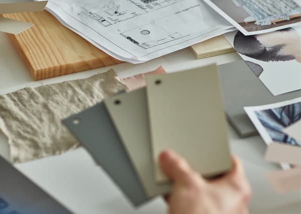 Une main tenant des échantillons de couleur dans les tons gris et beige, au-dessus d’un bureau couvert de plans, photos et échantillons.