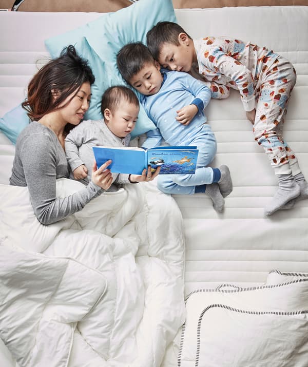 Une maman et ses trois enfants bien installés dans un lit tout blanc, en pleine lecture d’une histoire.