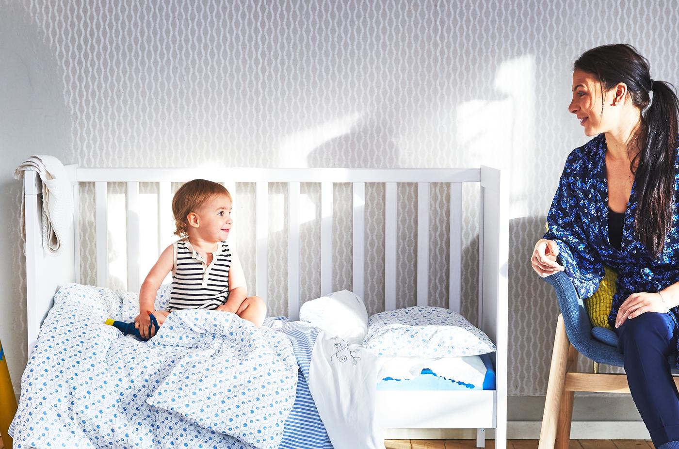Une mère, assise sur une chaise en bois, et son bébé, assis dans un lit à barreaux blanc ouvert sur un côté et couvert de textiles à motifs bleus, se regardent.