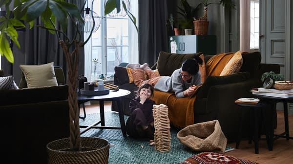 Une mère sur un canapé FÄRLÖV et son enfant sur le plancher jouent ensemble dans un salon où se trouvent des plantes, des rideaux et des tables basses.