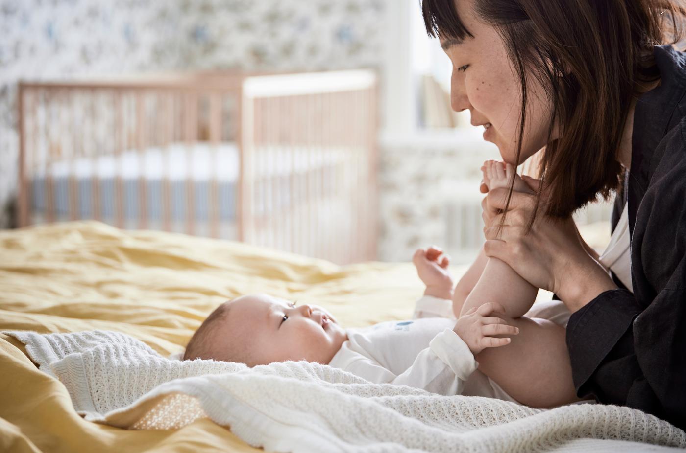 Une mère tient les orteils de son bébé et sourit, tandis que le poupon est couché sur des draps jaunes, avec un lit à barreaux en arrière-plan.