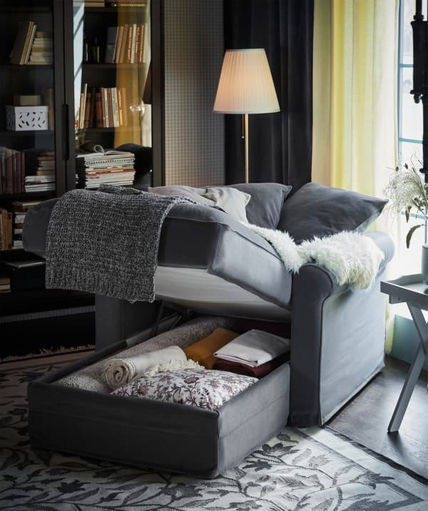 Une méridienne grise GRÖNLID avec l’espace de rangement intégré sous l’assise qui laisse entrevoir des plaids et des coussins de réserve.