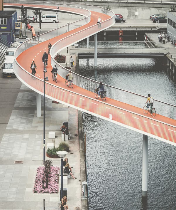 Une passerelle cyclable orange serpente au-dessus d'un cours d'eau et d'une rive aménagée pour les piétons.