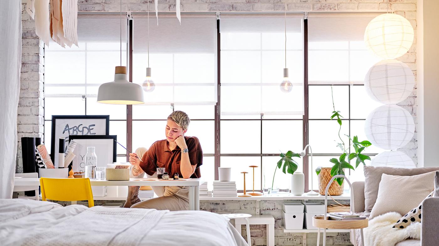 Une personne assise à un bureau LAGKAPTEN/ADILS blanc sous une suspension BUNKEFLO avec une table d’appoint LACK blanche derrière elle.