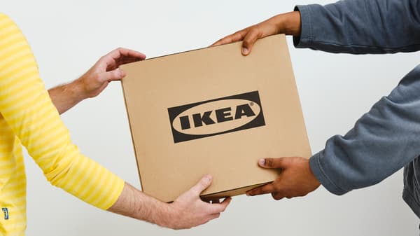 Une personne en pull gris donne une boîte en carton carrée avec un logo IKEA noir à une autre personne portant un haut de collaborateur IKEA jaune.