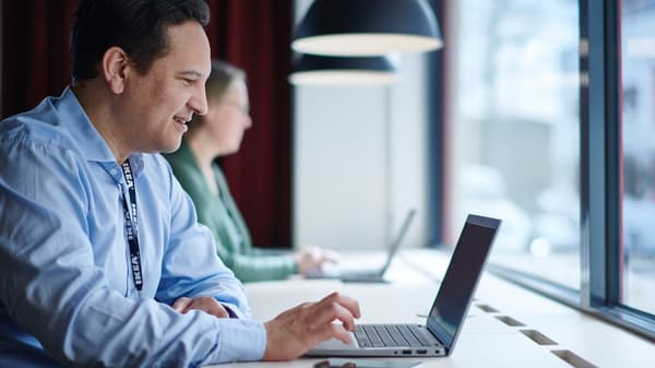 Une personne est assise à un bureau devant un ordinateur portable ouvert, face à une grande fenêtre.
