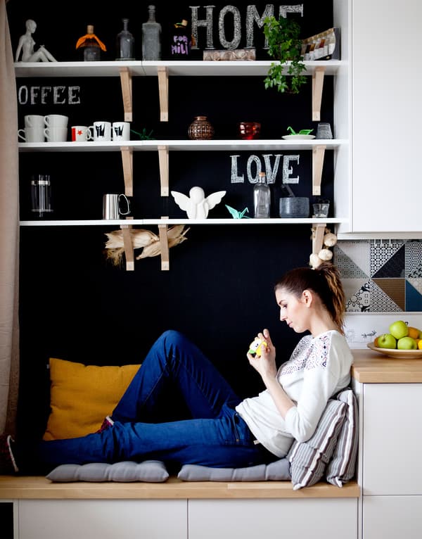 Une personne est assise sur un banc de rangement recouvert de coussins dans cette cuisine avec des armoires blanches et un mur noir.
