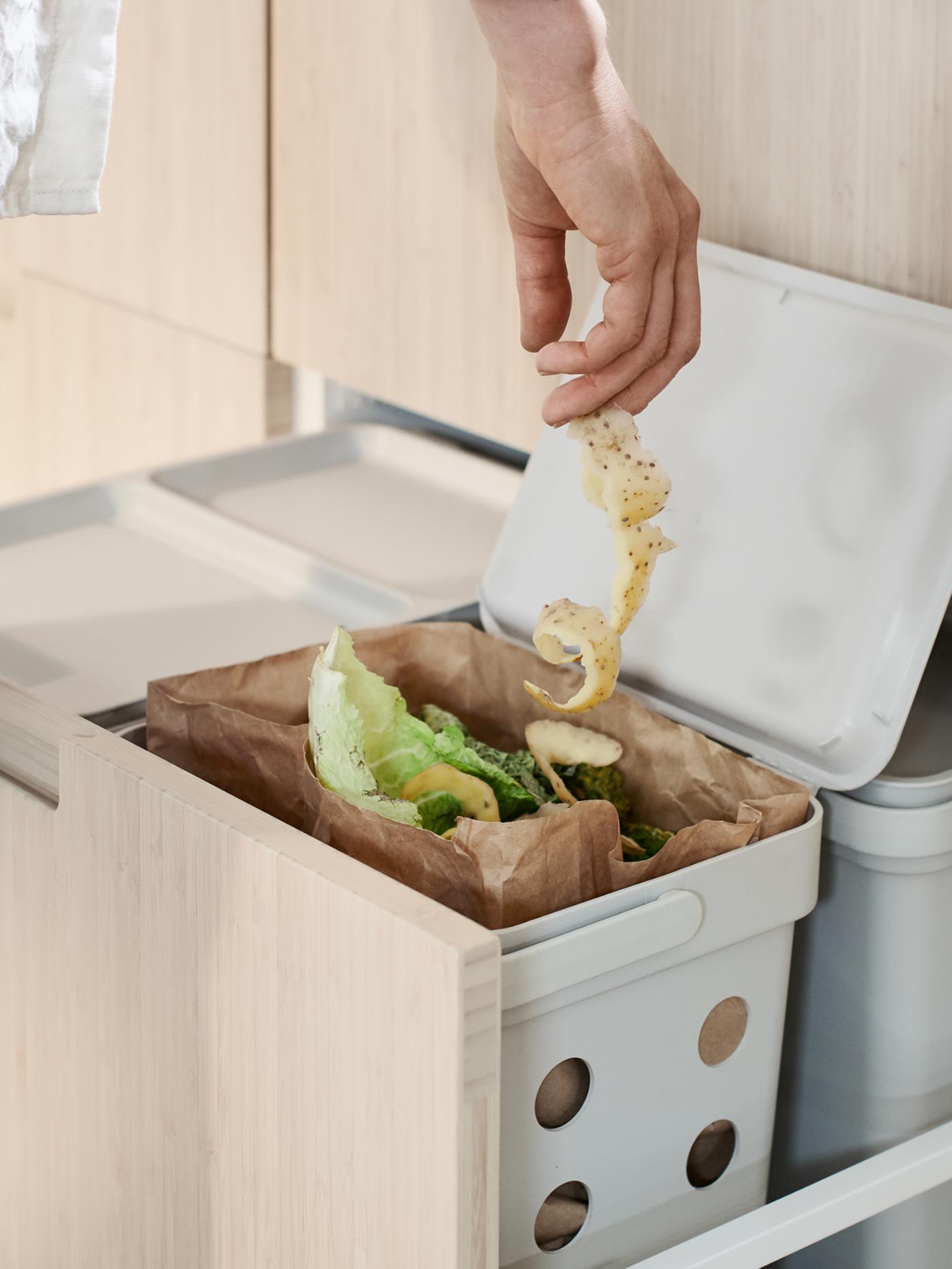 Une personne jette une épluchure de légume dans un sac à déchets alimentaires en papier brun. Cette dernière se trouve dans une solution de tri des déchets HÅLLBAR qui, elle, est installée dans un tiroir haut MAXIMERA.