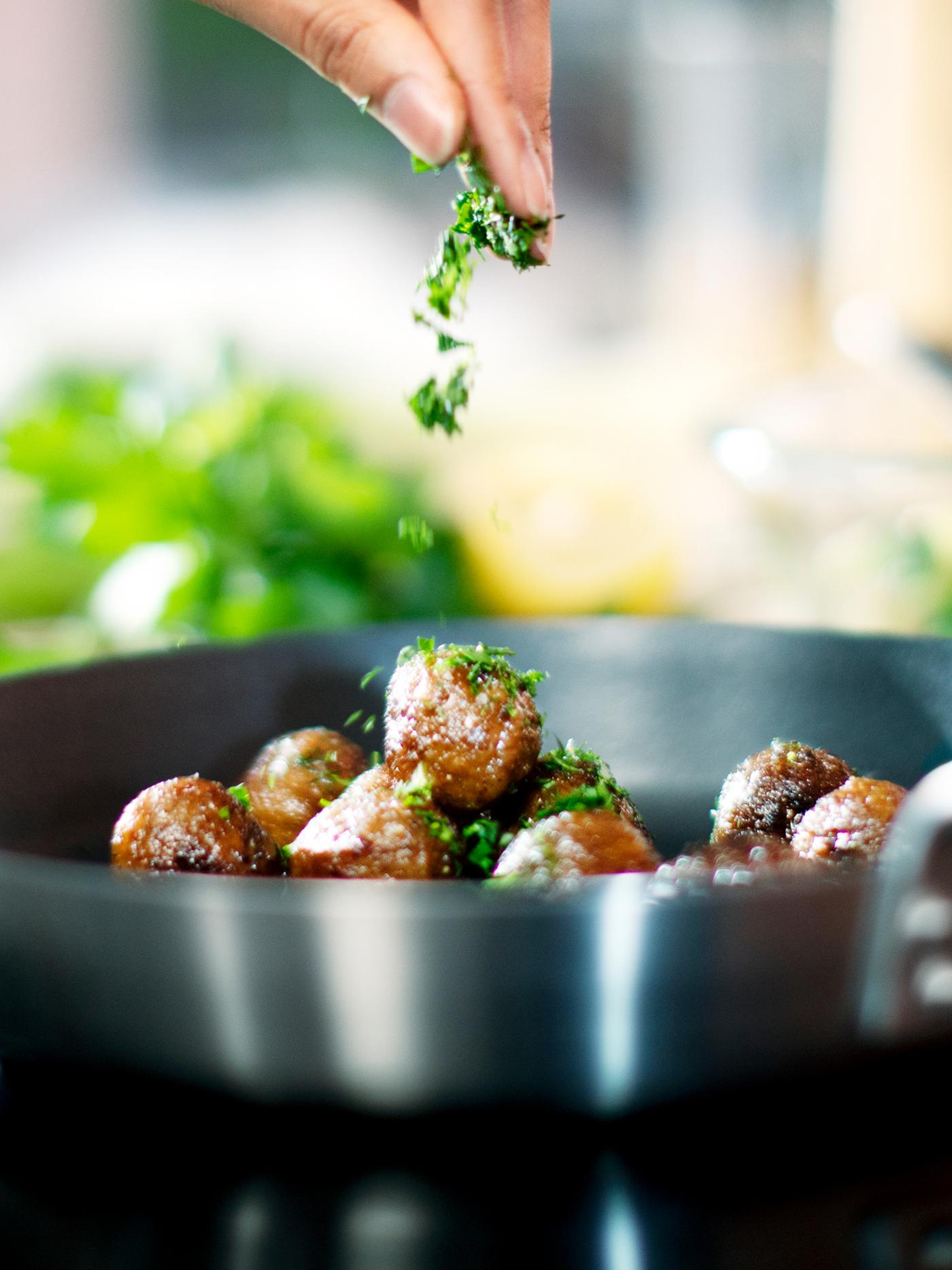 Une personne saupoudre des herbes vertes hachées sur des boulettes végétales HUVUDROLL disposées dans une poêle IKEA 365+ placée sur une table de cuisson.
