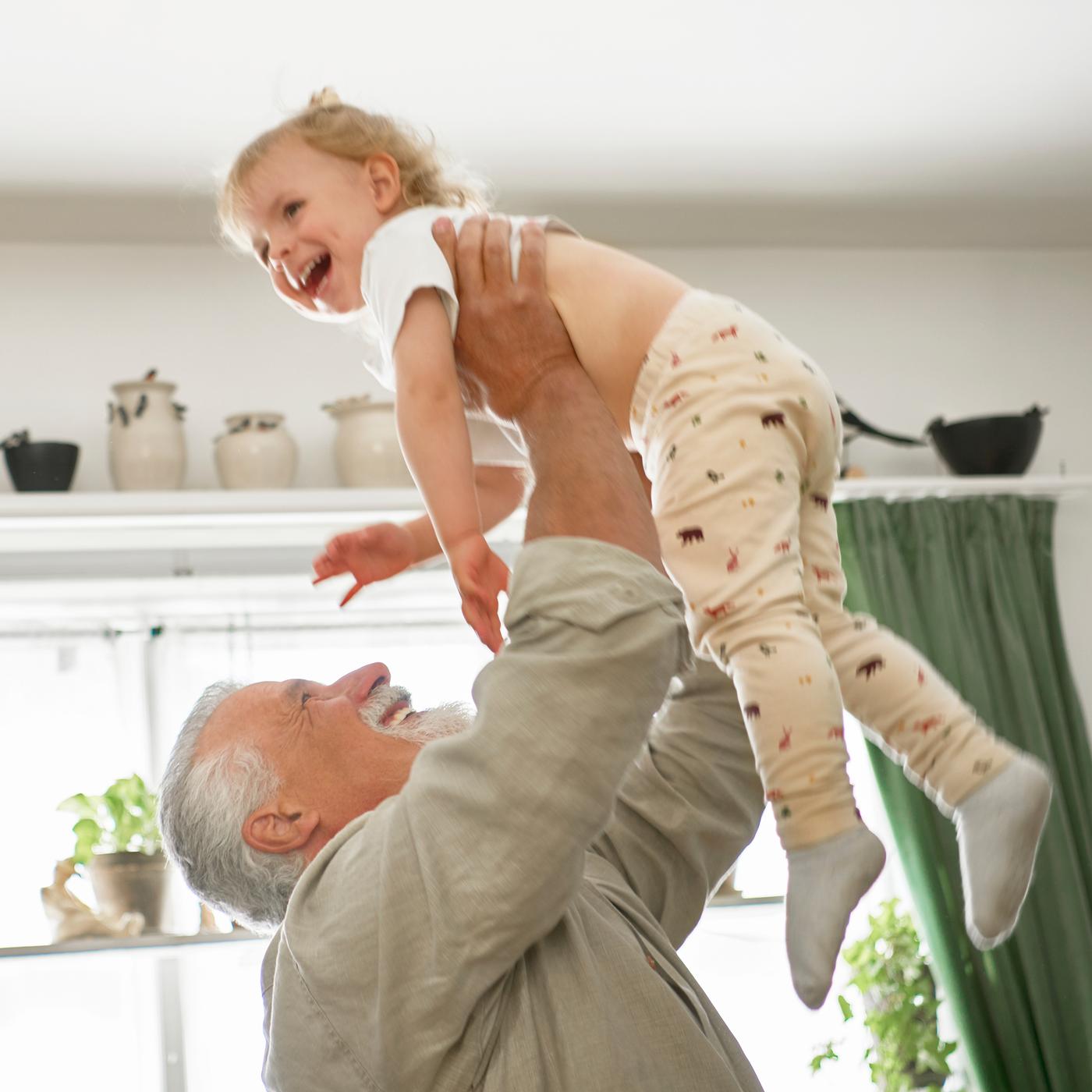 Une personne souriante, barbe et cheveux gris, soulève à bout de bras au-dessus de sa tête un enfant souriant, sous un plafond blanc.