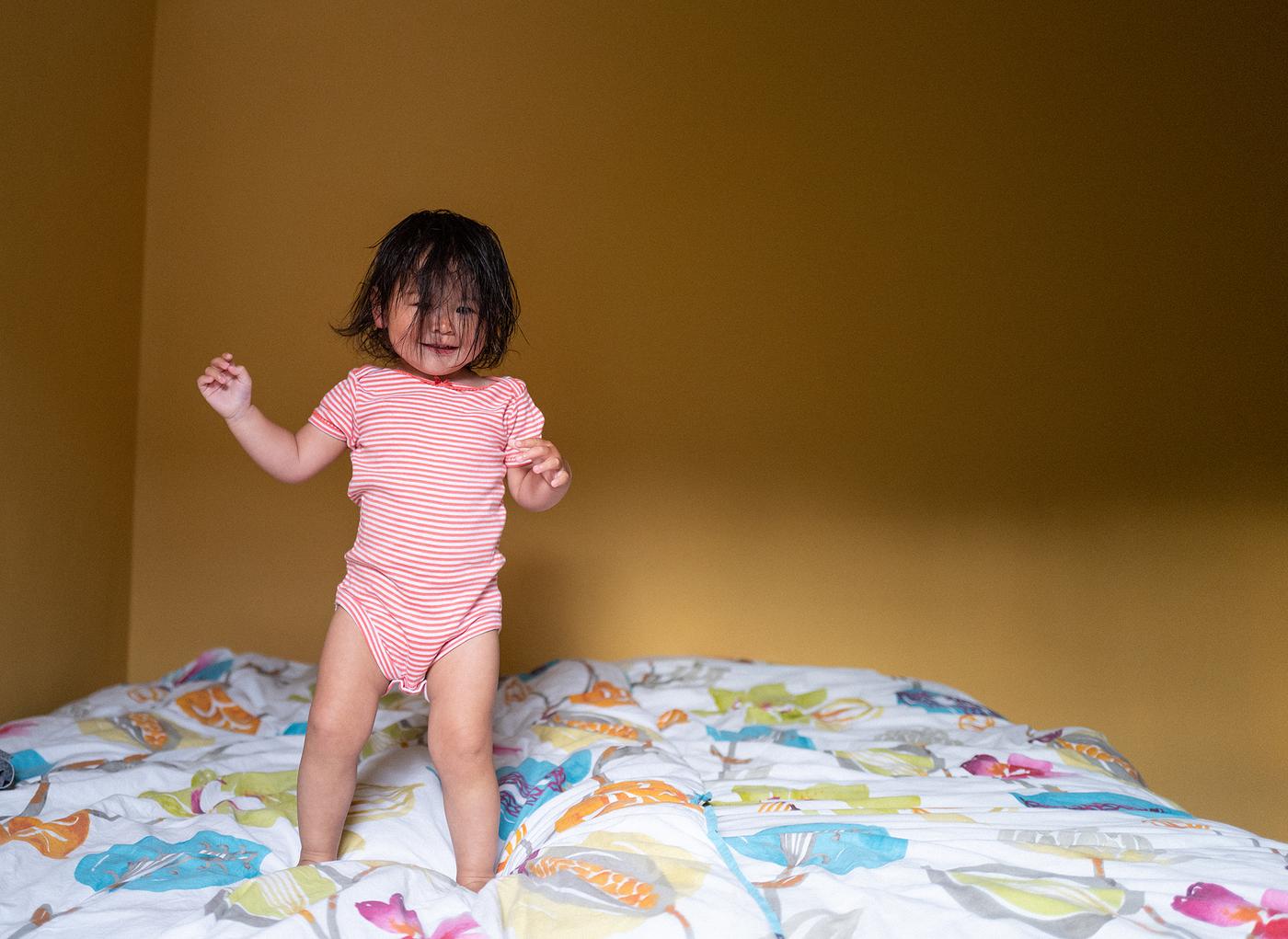 Une petite fille aux cheveux foncés couvrant son visage, sautant joyeusement sur un lit.