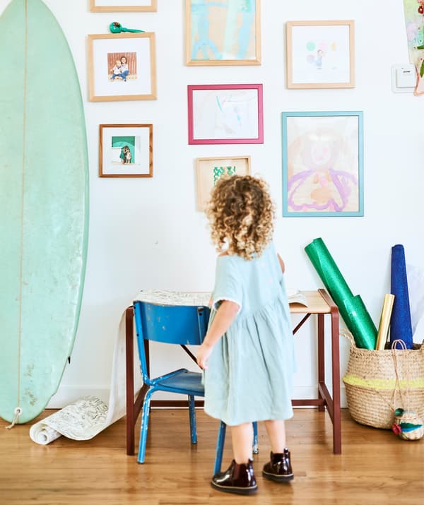 Une petite fille devant un petit bureau avec chaise, regardant le mur où sont accrochés peintures et photos de famille, à côté d'une planche de surf.