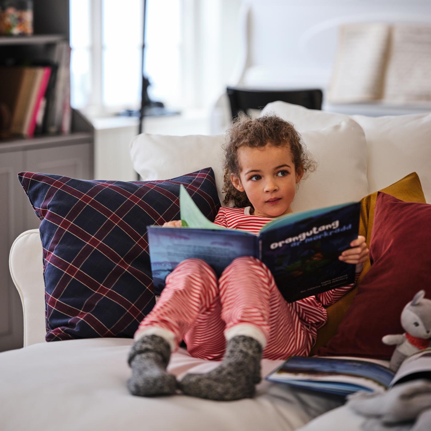 Une petite fille en pyjamas, confortablement installée avec un libre dans un canapé 4 places avec méridienne GRÖNLID blanc.