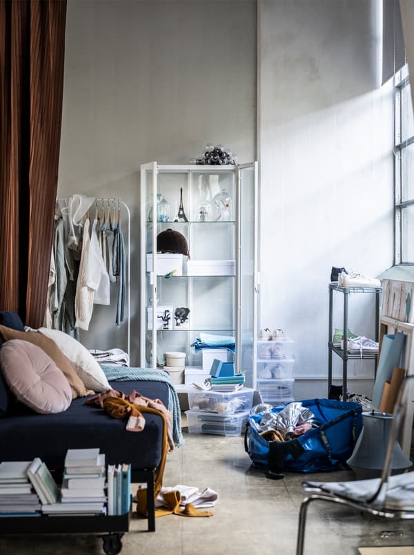 Une pièce en désordre avec un canapé, une étagère blanche, des rideaux, des livres empilés et un sac bleu IKEA rempli d’affaires posé sur le sol. La lumière du jour entre par la fenêtre.