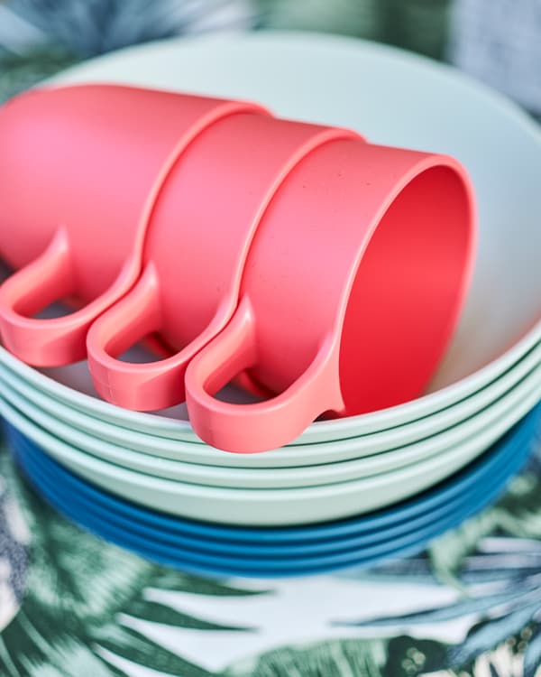 Une pile d’assiettes bleu foncé et bleu clair avec trois mugs rouge clair empilés dessus, sur un tissu à imprimé de feuilles bleues et vertes.