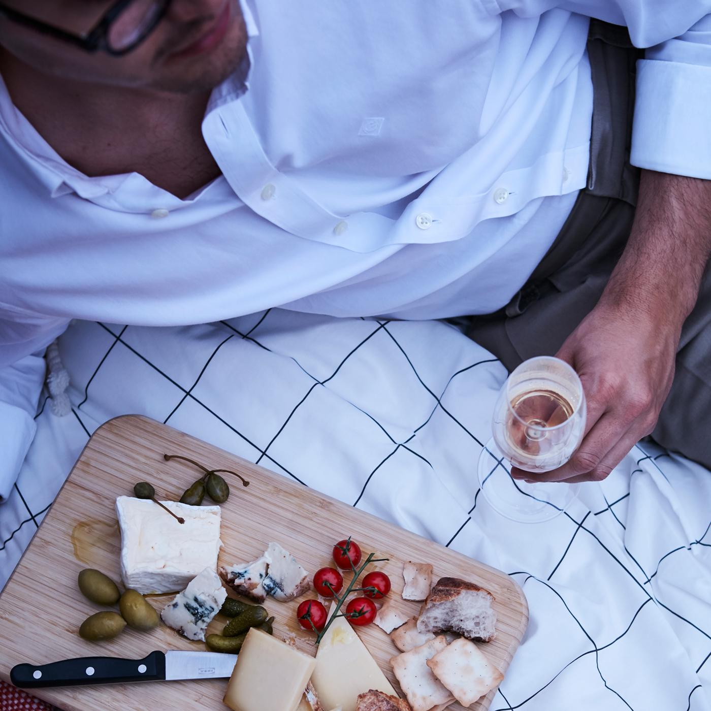 Une planche à découper en bois avec des hors-d’œuvre comme des fromages, des olives et des craquelins placés sur une housse de couette avec un homme à côté.