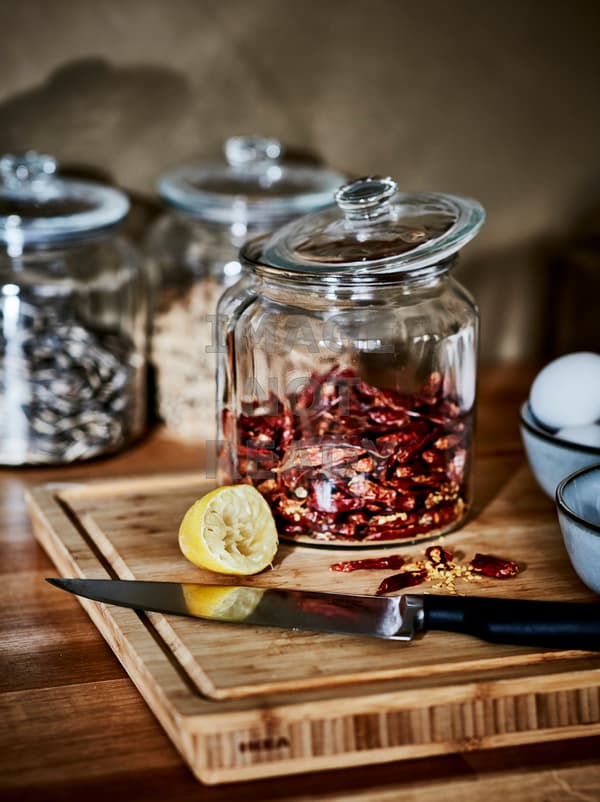 Une planche à découper en bois avec un couteau, un citron pressé et trois bocaux en verre VARDAGEN remplis de graines et de piment.
