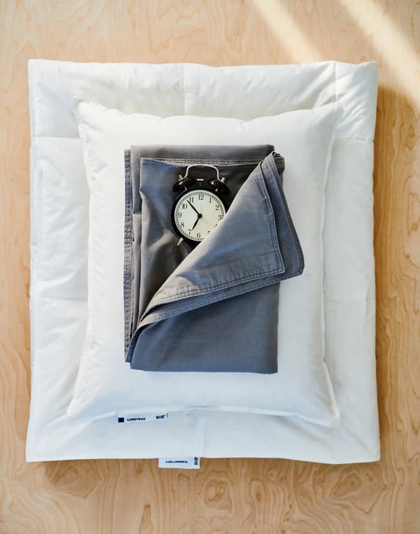 Une plaque de contreplaqué avec une couette blanche pliée, un oreiller blanc, du linge de lit bleu foncé et un réveil à l’ancienne noir.