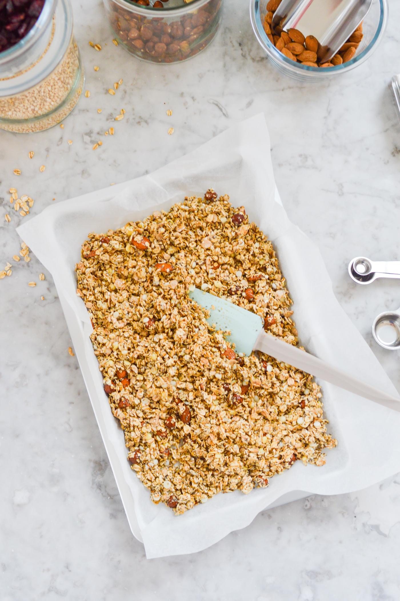 Une plaque de cuisson recouverte de muesli non cuit sur du papier sulfurisé, posée sur un plan de travail en marbre, entourée de bocaux contenant les ingrédients et d'une spatule bleu clair.