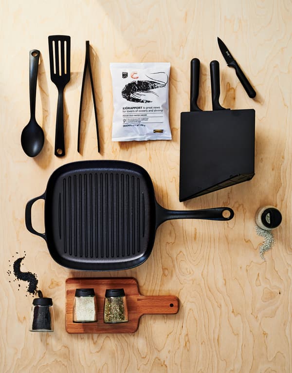 Une poêle en fonte VARDAGEN avec des ustensiles, un range-couteaux avec des couteaux, une planche à découper en bois, des épices et des crevettes congelées.