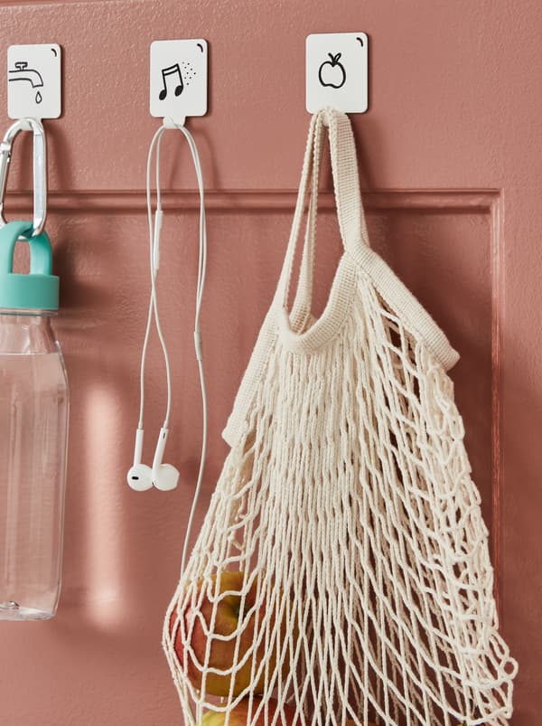 Une porte rose foncé dotée de crochets à motifs graphiques auxquels sont accrochés une bouteille d’eau, des écouteurs et un sac filet KUNGSFORS contenant des fruits.