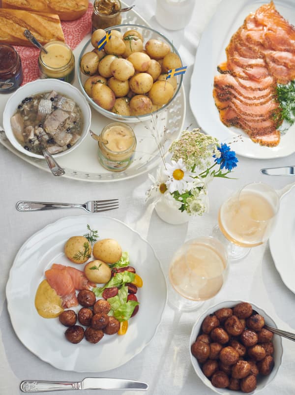 Une portion de table avec des assiettes, des couverts ÄTBART, du saumon mariné sur un plat, des boulettes de viande dans un bol et du hareng sur un plateau ROMANTISK.