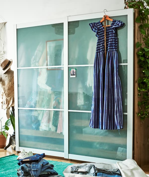 Une robe bleue est suspendue à l’avant d’une garde-robe à portes coulissantes, un mannequin de couture se trouve à côté, et devant, une pile de jeans.