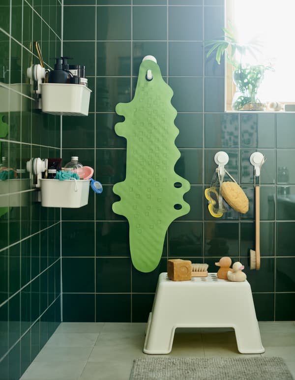 Une salle de bain avec un marchepied et un tapis de baignoire PATRULL en vert mat en forme de crocodile suspendu à un crochet avec ventouse.