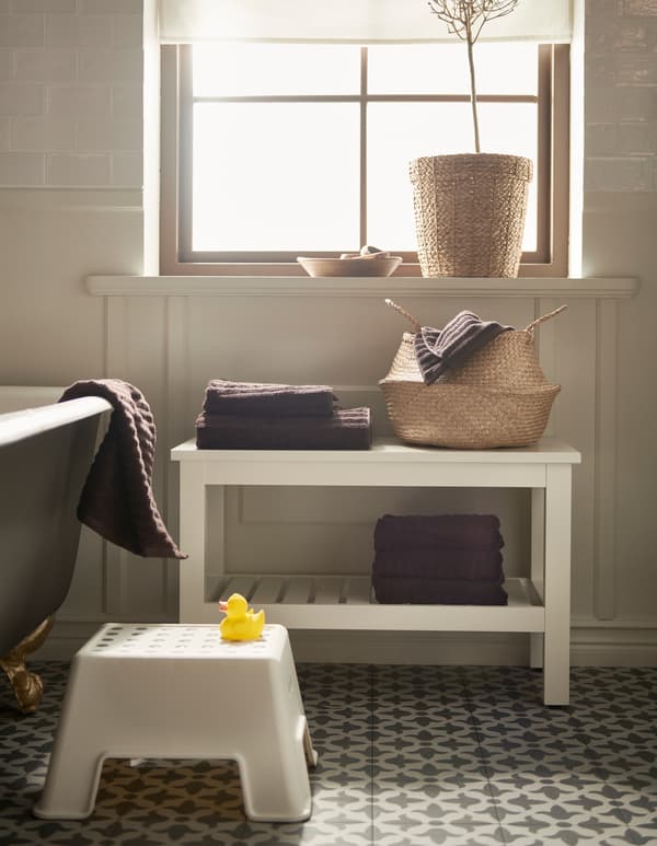 Une salle de bain avec un panier en rotin, un marchepied BOLMEN blanc et un banc HEMNES blanc avec des tablettes pour les serviettes.