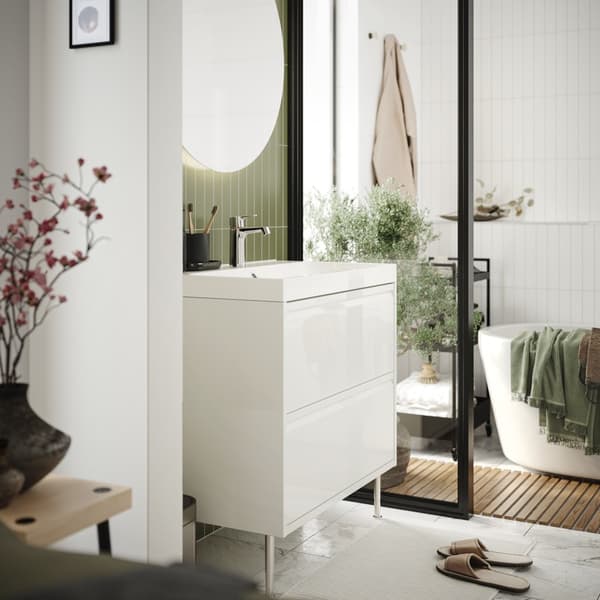 Une salle de bains avec un meuble sous-vasque IKEA ÄNGSJÖN blanc avec poignées intégrées, une baignoire derrière et une petite étagère de salle de bains à roulettes