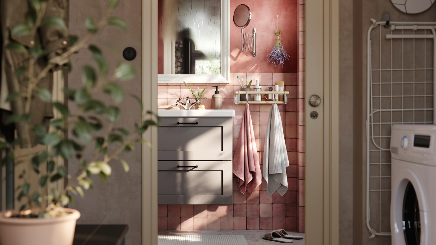 Une salle de bains carrelée rose meublée d’une armoire ENHET grise avec deux tiroirs, un miroir blanc et deux serviettes suspendues à un rail doté de crochets.
