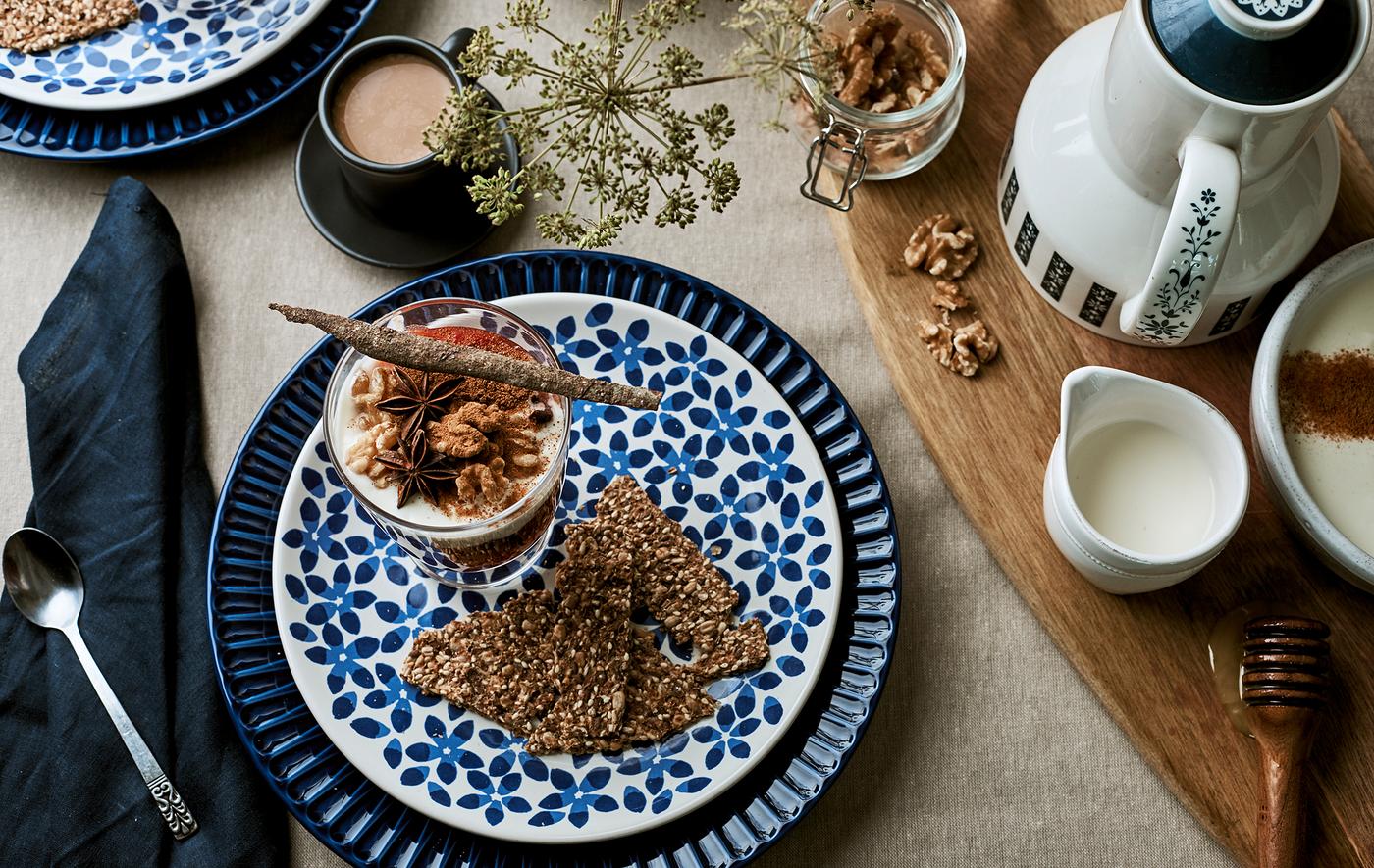 Une table de petit-déjeuner dressée avec une nappe en lin, des assiettes à motif bleues, du granola dans un verre et des accessoires pour le café sur une planche à découper.