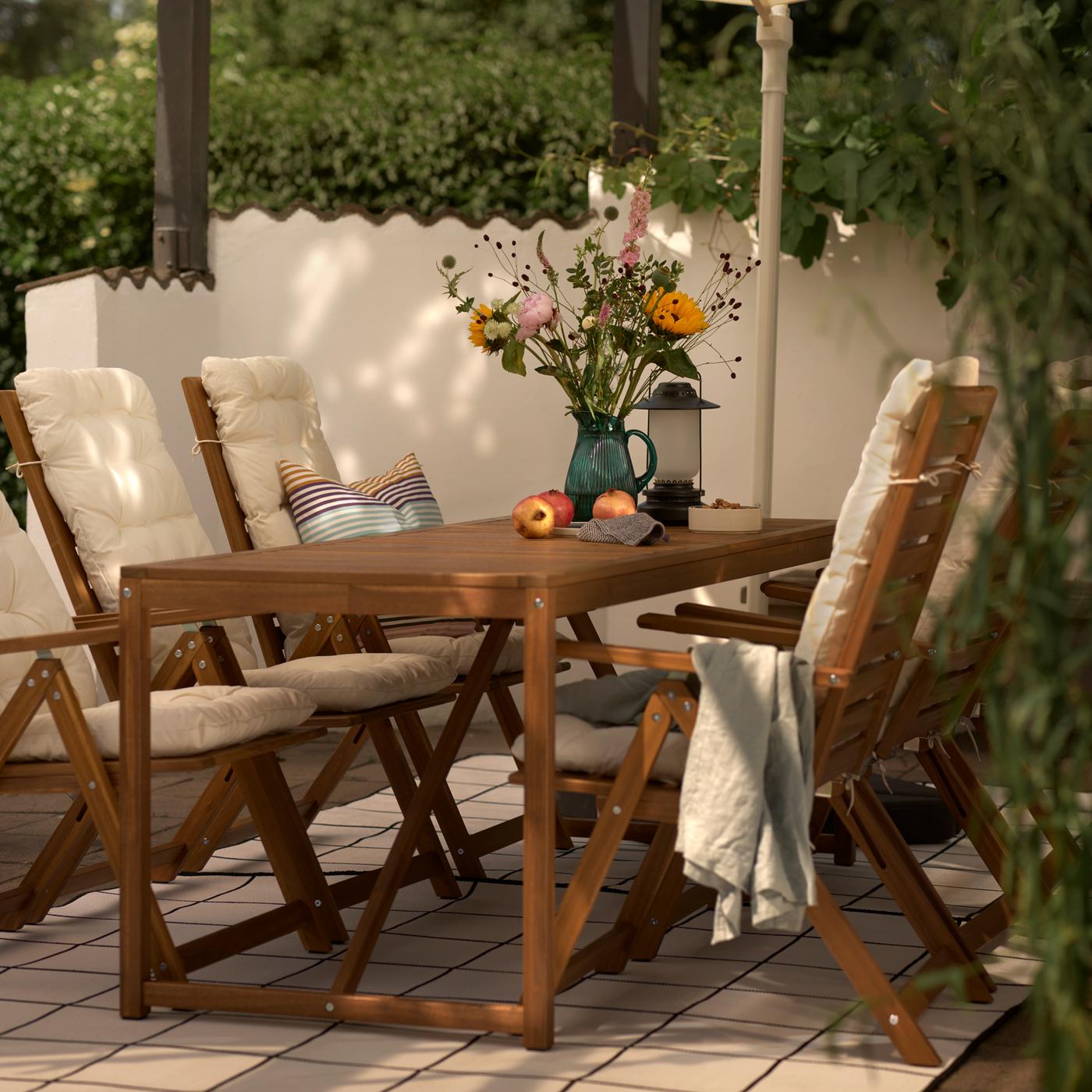 Une table d'extérieur brun clair avec des fleurs dans un pichet à motif/vert, plus six fauteuils à dossier inclinable garnis de coussins assise/dossier beiges.