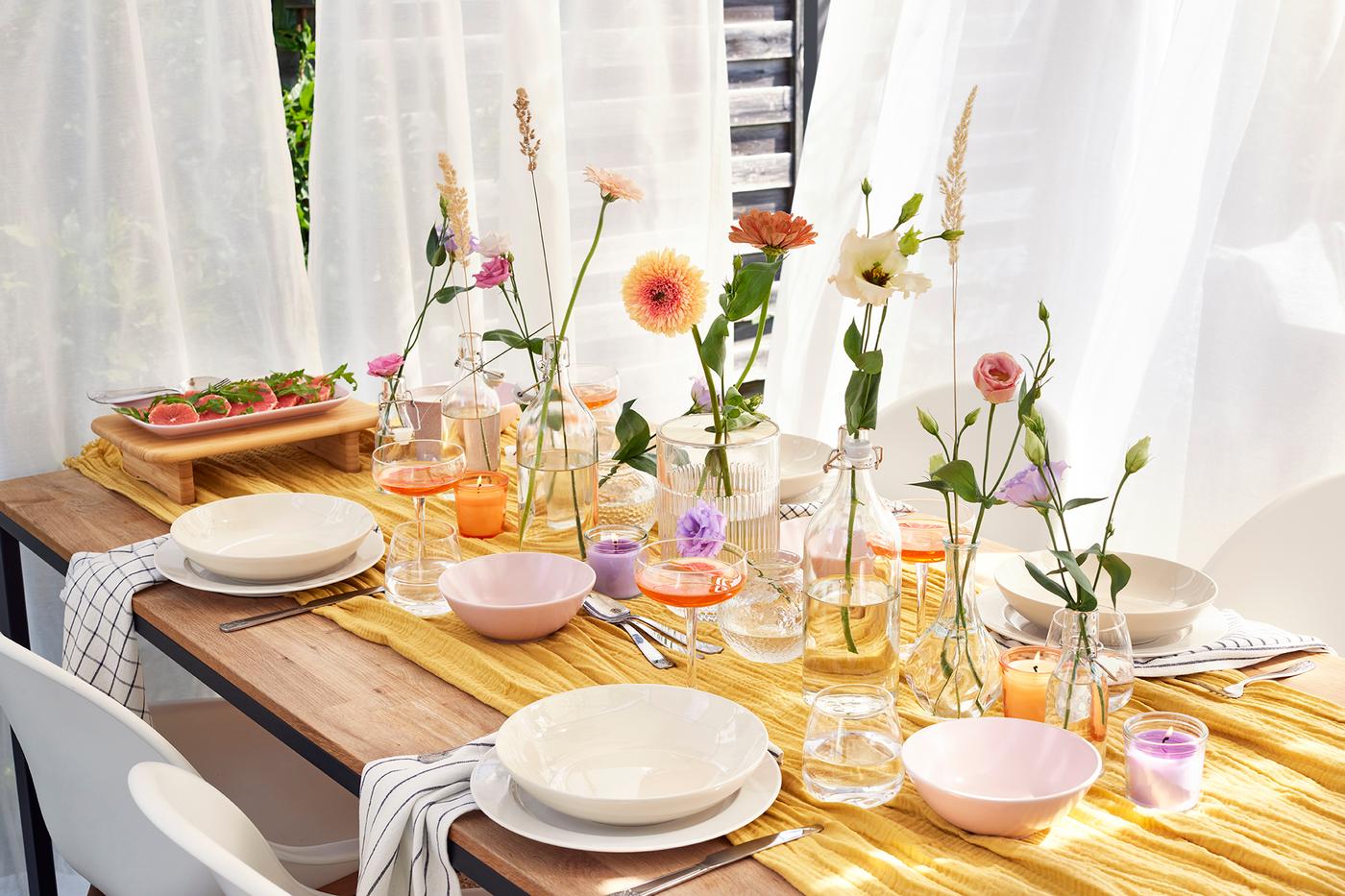 Une table dressée devant des rideaux blancs est couverte d'assiettes ,de bols et de plusieurs vases avec des fleurs et des herbes individuelles.