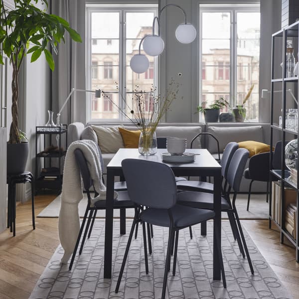 Une table noir/brun foncé, des chaises grises, un tapis à motifs géométriques, une suspension chromée/blanc et un canapé gris clair.