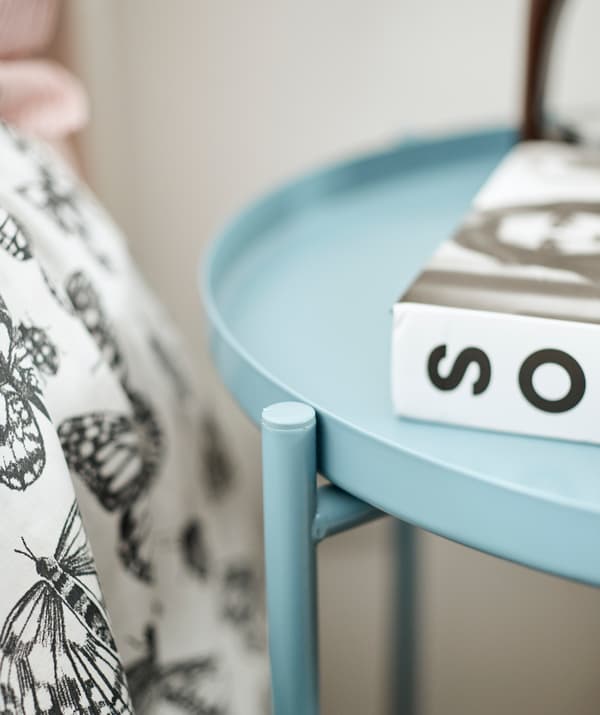 Une table ronde bleue en métal sur laquelle est posé un livre, à côté d’un lit revêtu d’une couette en noir et blanc.
