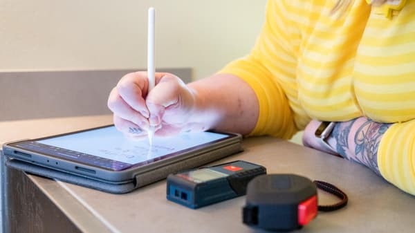 Une tablette sur laquelle quelqu'un dessine un plan de cuisine à l'aide d'un stylet. A côté se trouvent un mètre ruban et un appareil de mesure au laser. 