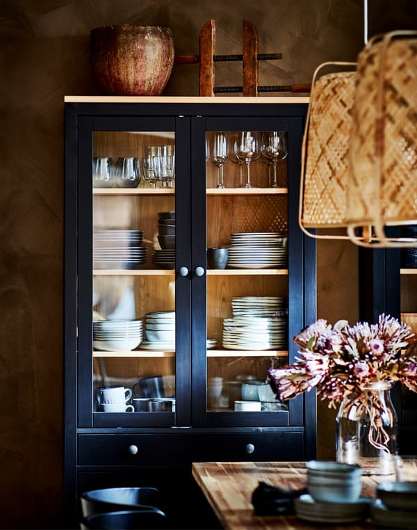 Une vitrine HEMNES au fini noir-brun avec verres à vin et vaisselle, placée derrière une table avec un vase à fleurs.