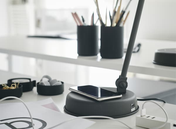 Uno smartphone in carica sulla base di una lampada da lavoro grigio scuro, su una scrivania bianca - IKEA