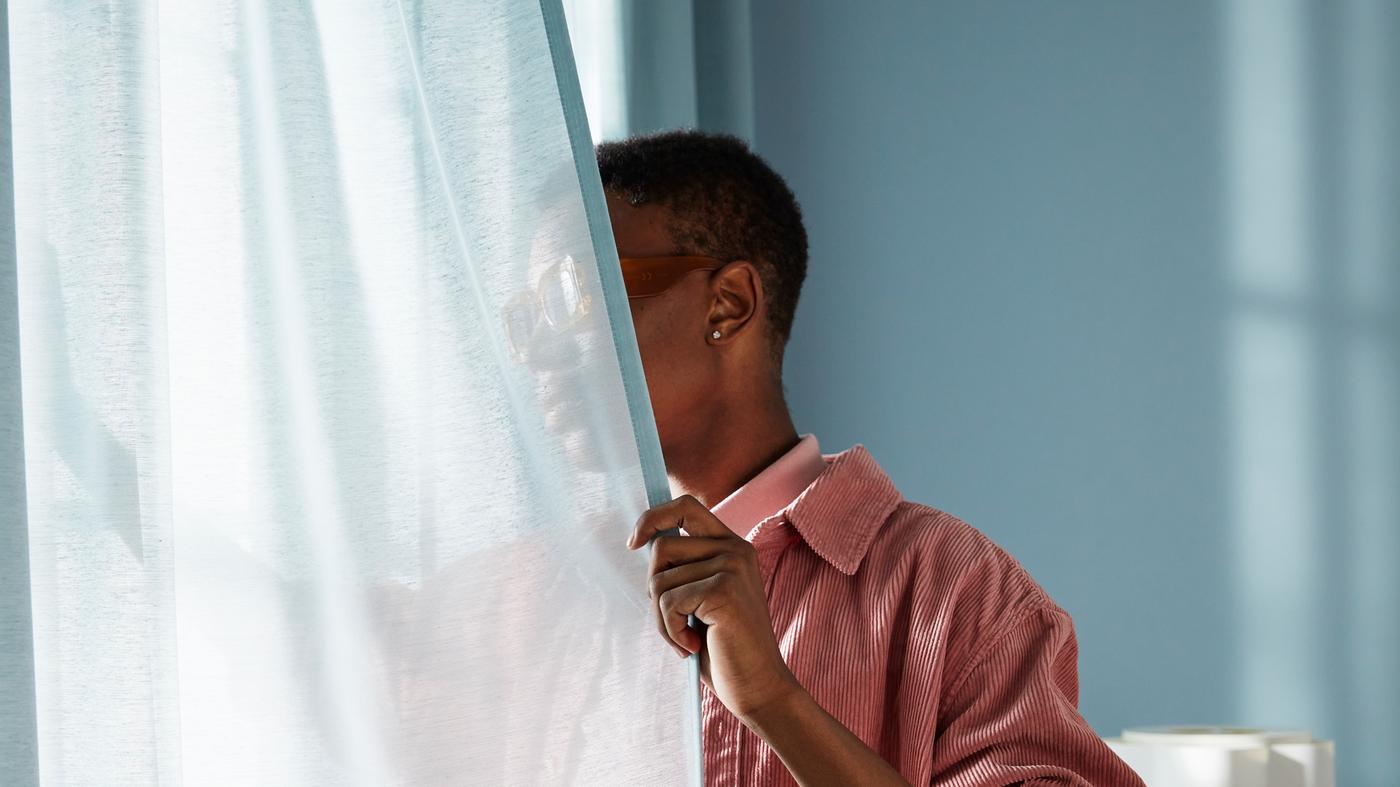 Uomo appoggiato su una tenda azzurra e che guarda fuori attraverso la finestra da una stanza azzurra.