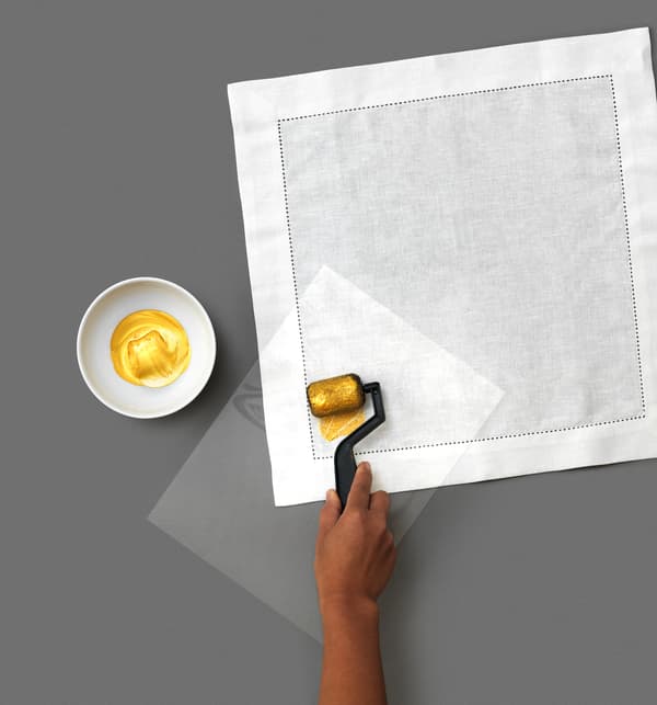 „Uradi sam” rešenje sjajno je ako želiš da napraviš ukrasne salvete po meri, koristeći bele GULLMAJ platnene salvete kompanije IKEA. Treći korak je da postaviš isečeni plastični šablon na salvetu. Zatim valjkom nanesi boju za tkaninu na salvetu.