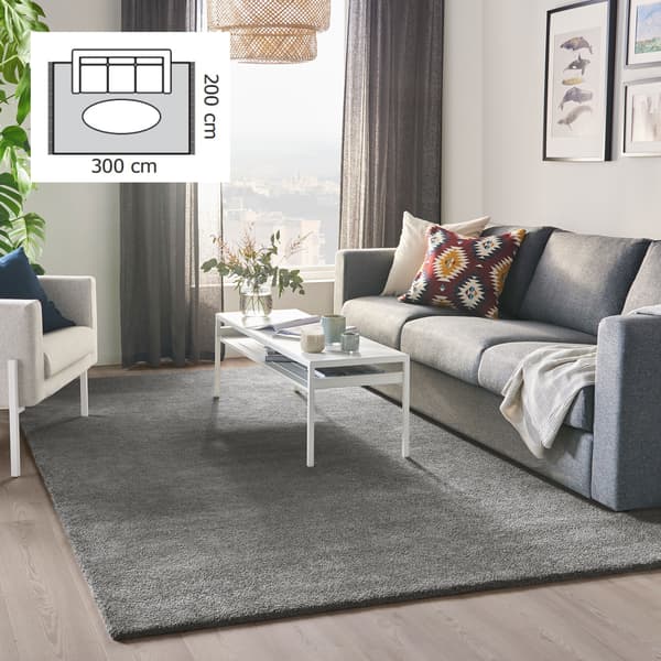 Vardagsrum med grå matta (300x200 cm), vitt soffbord, soffa med färgglada kuddar, fåtölj, växt och gardiner. Uppe i vänstra hörnet finns en liten planritning över ett vardagsrum som visar en matta i storleken 300x200 cm.