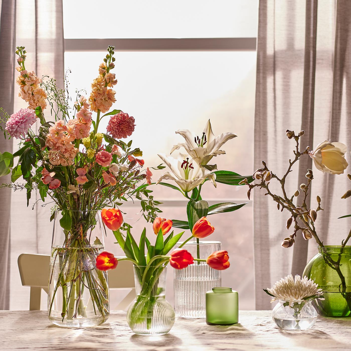 Vases in different sizes holding flowers, all placed on a table with a beige tablecloth in front of a sunlit window.