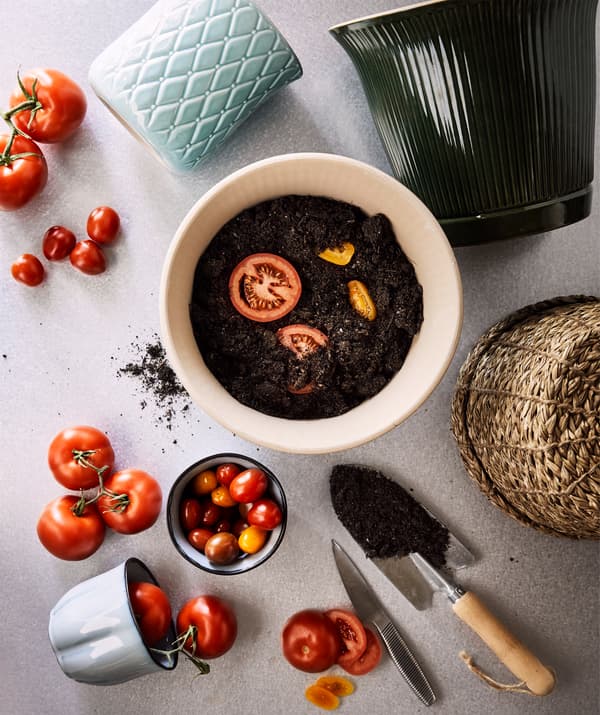 Verschiedene Übertöpfe, u. a. KAMOMILL Übertopf in Grün, reife Tomaten und Scheiben gelber und roter Tomaten, die in frische Erde gelegt wurden.
