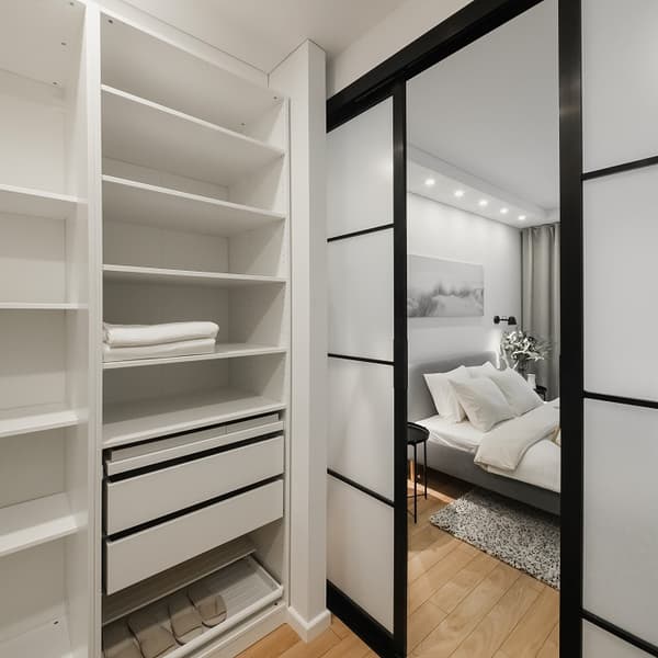 View from a walk-in closet into a cozy, modern bedroom through black-framed sliding glass doors. The closet features white open shelving with folded linens, organized drawers, and storage boxes on the bottom shelf. The bedroom beyond has a neatly made bed with soft pillows, ambient ceiling lighting, and minimalist decor.