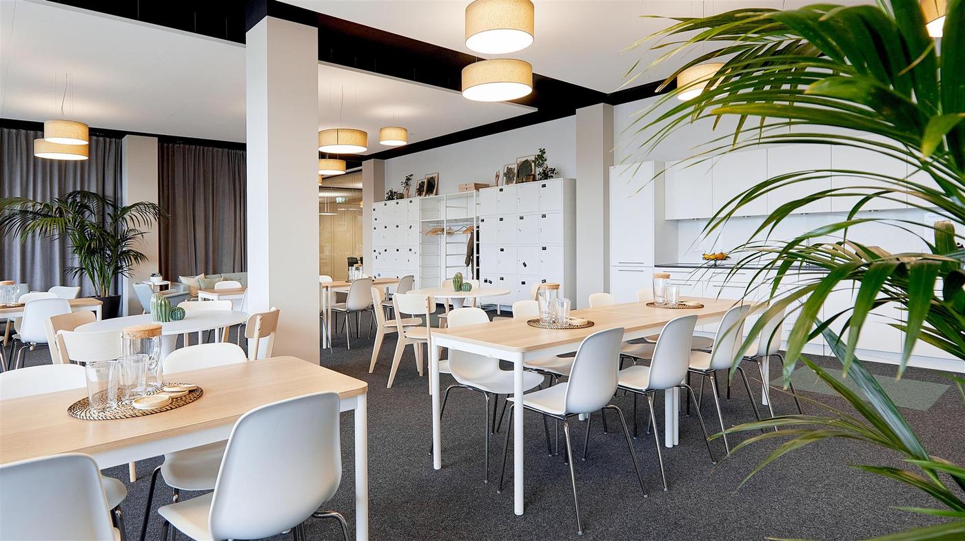 View into an office kitchen with white kitchen units and dining tables with carafes and glasses on top. 