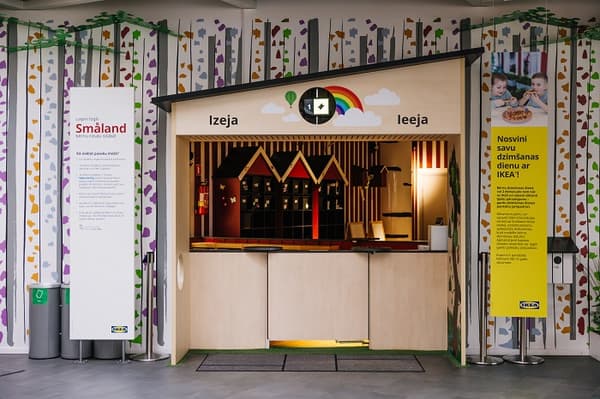 Вход и выход из игровой зоны IKEA Småland, оформленной в виде небольшого домика с радужной вывеской, красочными настенными рисунками на лесную тематику и информационными табличками на латышском языке, приглашающими детей поиграть и отпраздновать свои дни рождения.