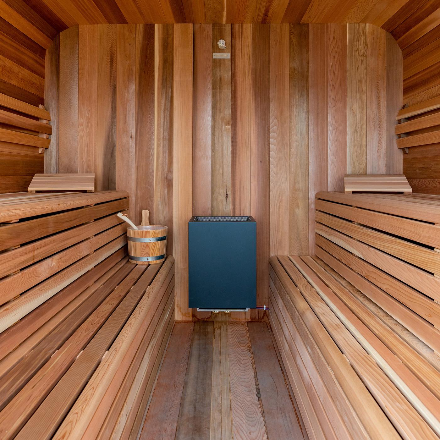 Vue à l’intérieur d’un « Sauna-Cube » aménagé en bois clair avec des bancs à plusieurs niveaux des deux côtés. Le four du sauna se trouve contre la cloison arrière